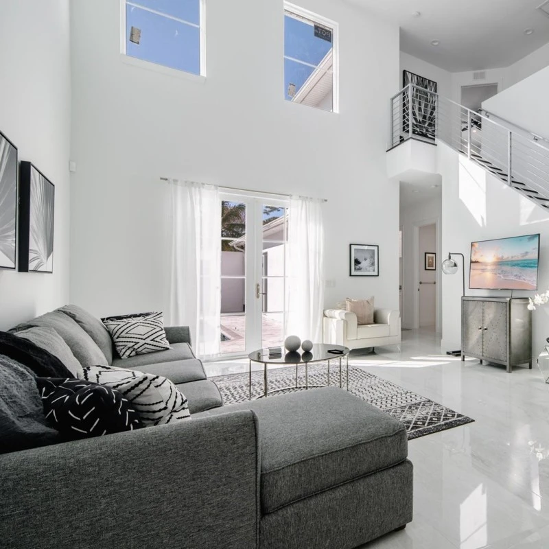 a well lit living room that is white with a gray couch, a table in the middle, many paintings on the wall, and a tv